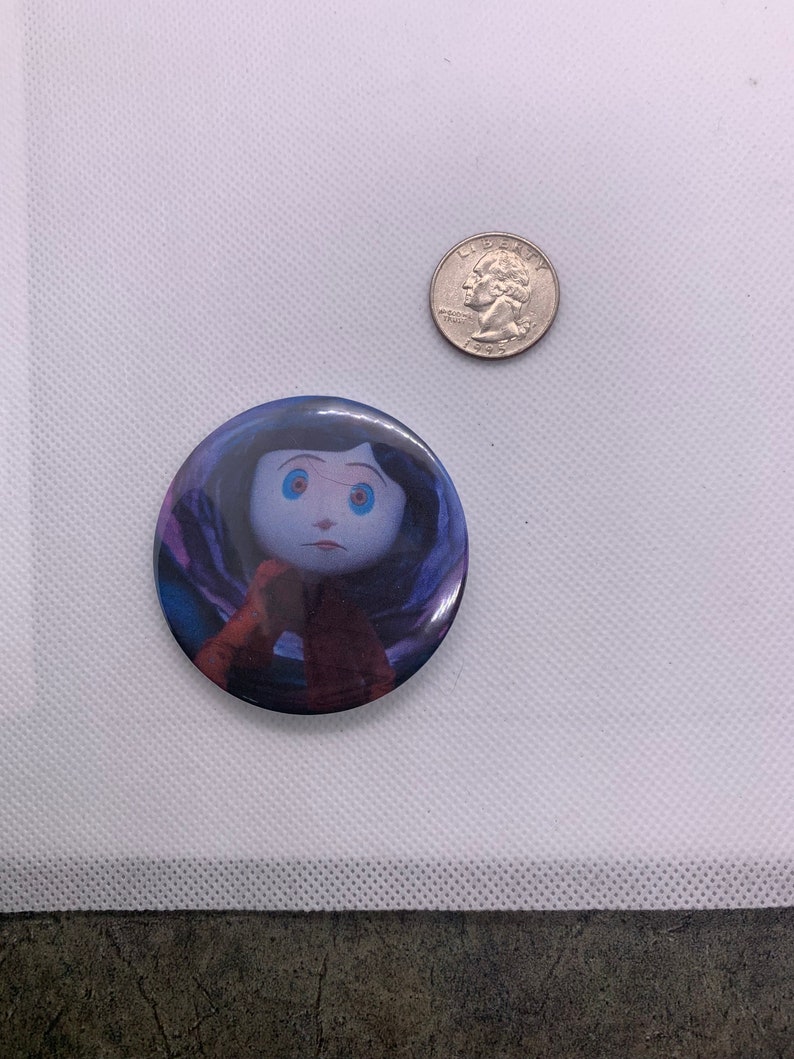 K&ouml;nnte beinhalten: Ein runder Knopf mit einem Bild eines M&auml;dchens mit blauen Augen und einem lila Kleid. Das M&auml;dchen schaut mit einem besorgten Ausdruck nach oben. Der Knopf ist etwa so gro&szlig; wie eine 25-Cent-M&uuml;nze.