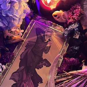 Op de afbeelding: Een ingelijste tarotkaart met een man in een donker pak en een halo. De kaart is omringd door gedroogde bloemen, een schedel en een kaars.
