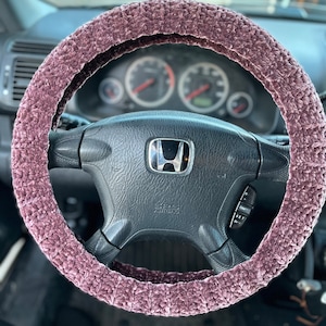 May include: A mauve, plush steering wheel cover is shown on a black steering wheel. The cover is made of a soft, textured material and fits snugly around the wheel. The car's dashboard and gauges are visible in the background.