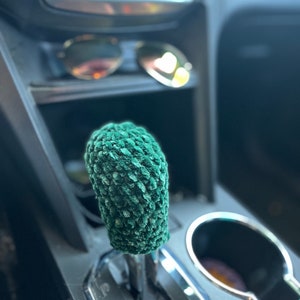 Peut inclure: Un couvre-pommeau de levier de vitesses vert et texturé, crocheté. Le couvre-pommeau est cylindrique et s'adapte au levier de vitesses d'une voiture. L'arrière-plan montre l'intérieur de la voiture, y compris le tableau de bord et un porte-gobelet.