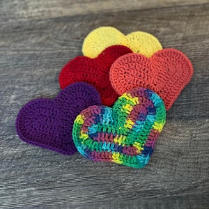 Puede incluir: Una colección de seis objetos en forma de corazón hechos a ganchillo en varios colores, incluyendo morado, rojo, amarillo y un corazón multicolor. Los corazones están dispuestos sobre una superficie de madera, mostrando la textura hecha a mano y los colores vibrantes.