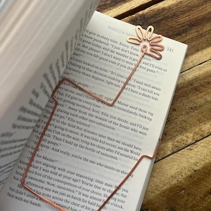 May include: A copper-colored metal bookmark in the shape of a flower is inserted into a book. The bookmark is holding the pages open. The book is open to page 145, with text visible.