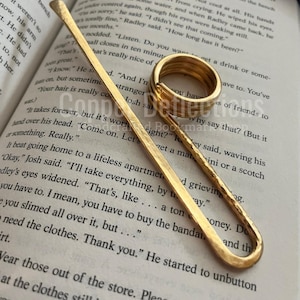 Puede incluir: Dos marcap&aacute;ginas de metal dorado. Uno es un marcap&aacute;ginas largo, delgado y recto. El otro es un marcap&aacute;ginas m&aacute;s grueso, redondeado y con un bucle abierto. Ambos marcap&aacute;ginas est&aacute;n sobre un libro con texto.