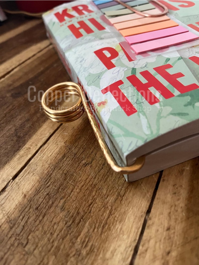 Puede incluir: Un sujetalibros de metal dorado con un dise&ntilde;o en espiral. El sujetalibros sostiene un libro con un patr&oacute;n floral verde y blanco. El libro tiene las palabras "KR HI" y "PLAY THE" impresas en la portada.