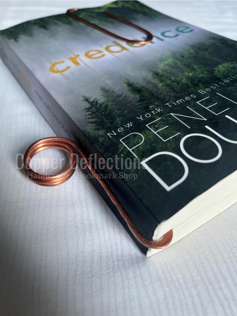 May include: A copper wire bookmark with a curled design. The bookmark is on top of a book titled "Credence" by Penelope Douglas. The book cover features a forest scene with the text "New York Times Bestseller" on the front.