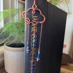 May include: A copper-colored cloud-shaped bookmark with dangling chains and blue beads. The bookmark is placed in a black book with the text "JACKIE COLLINS HOLLYWOOD" visible on the spine. The background includes greenery and a copper-colored mirror.