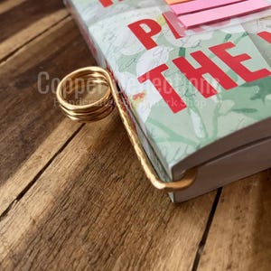 May include: A gold metal book holder with a spiral design. The book holder is holding a book with a green and white floral pattern. The book has the words "KR HI" and "PLAY THE" printed on the cover.