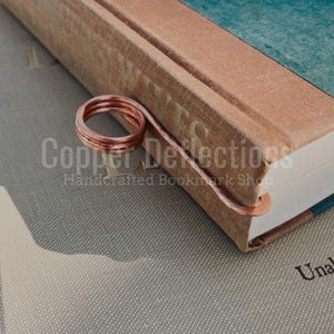 May include: A copper wire bookmark in the shape of a spiral, resting on the spine of a book titled "Grey Wives".