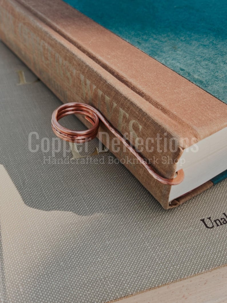 May include: A copper wire bookmark in the shape of a spiral, resting on the spine of a book titled "Grey Wives".