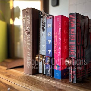 May include: A set of bookends made of copper wire, holding up a collection of books with various titles. The bookends are shaped like a pair of rings, and the books are all different colors and sizes.