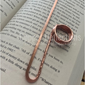 Puede incluir: Dos anillos de cobre con las palabras "Angela" y "He's that good guy b" grabadas en ellos. Los anillos est&aacute;n sobre un libro con texto sobre un anillo de boda.