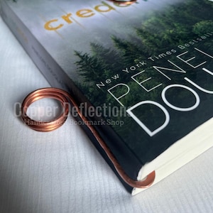 Puede incluir: Un marcap&aacute;ginas de alambre de cobre con un dise&ntilde;o rizado. El marcap&aacute;ginas est&aacute; encima de un libro titulado "Credence" de Penelope Douglas. La portada del libro presenta una escena de bosque con el texto "New York Times Bestseller" en la portada.