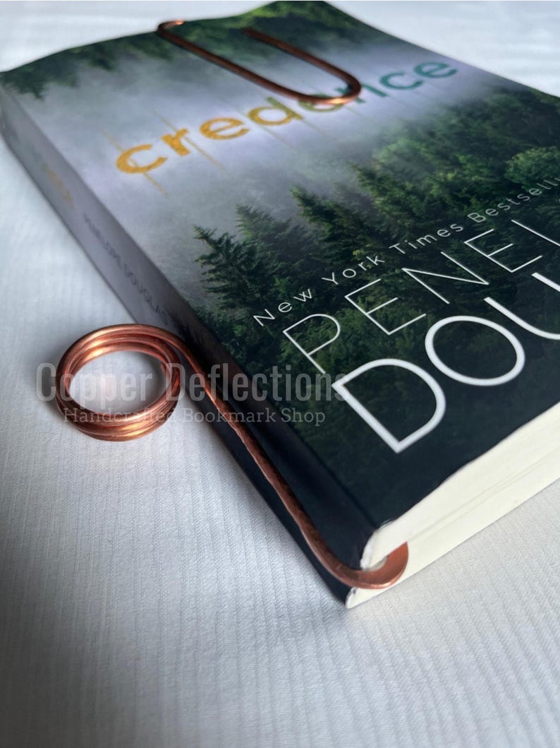 Puede incluir: Un marcap&aacute;ginas de alambre de cobre con un dise&ntilde;o rizado. El marcap&aacute;ginas est&aacute; encima de un libro titulado "Credence" de Penelope Douglas. La portada del libro presenta una escena de bosque con el texto "New York Times Bestseller" en la portada.