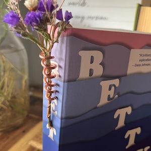 May include: A book titled "Betty" with a copper wire bookmark holding a bouquet of dried flowers. The book cover features a gradient of purple, blue, and pink colors with the title in white.