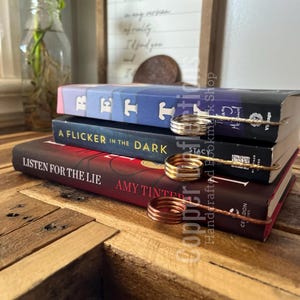 May include: Three books stacked on a wooden table with a copper wire bookmark holding the pages. The books are titled "Rett", "A Flicker in the Dark", and "Listen for the Lie".