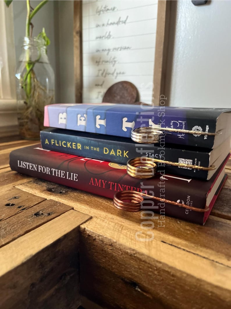 May include: Three books stacked on a wooden table with a copper wire bookmark holding the pages. The books are titled "Rett", "A Flicker in the Dark", and "Listen for the Lie".