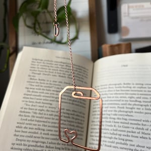 May include: A rose gold metal bookmark shaped like a book with a heart cutout. The bookmark is hanging from a chain.