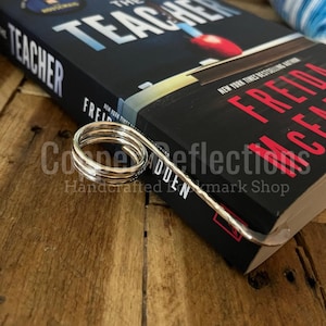 Puede incluir: Un marcap&aacute;ginas de metal plateado con un dise&ntilde;o de resorte. El marcap&aacute;ginas est&aacute; encima de un libro titulado "The Teacher" de Fredrick Backman. El libro est&aacute; sobre una superficie de madera.