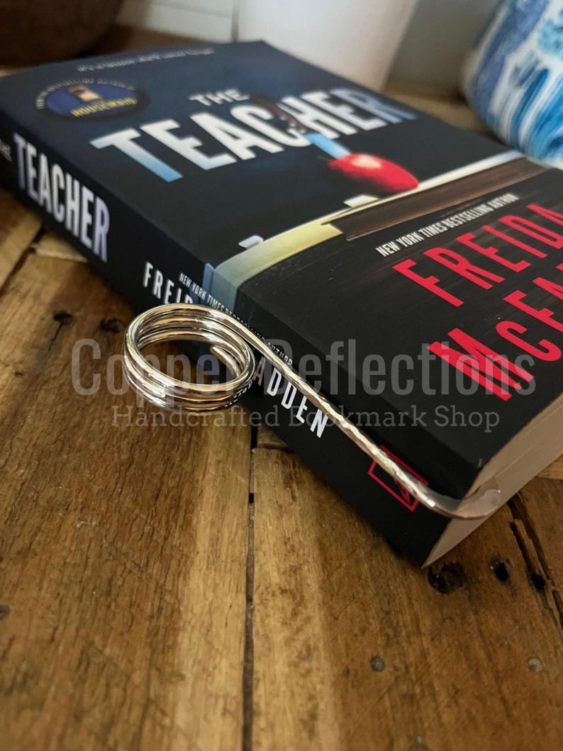 Puede incluir: Un marcap&aacute;ginas de metal plateado con un dise&ntilde;o de resorte. El marcap&aacute;ginas est&aacute; encima de un libro titulado "The Teacher" de Fredrick Backman. El libro est&aacute; sobre una superficie de madera.