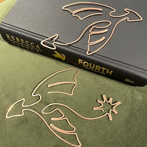 May include: Two gold-colored dragon-shaped bookmark designs. The top bookmark is on a black book with the text "REBECCA YARROS FOURTH WINE". The bottom bookmark is on a green surface.