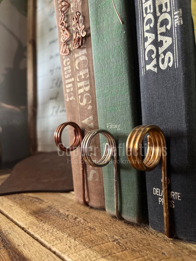 Puede incluir: Tres sujetalibros hechos de alambre met&aacute;lico en espiral. Los sujetalibros son de diferentes colores: cobre, plata y oro. Los sujetalibros sostienen una pila de libros con los t&iacute;tulos "The Icers", "Viking" y "The Egas Legacy".