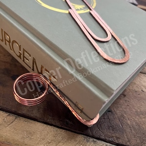 Puede incluir: Un marcap&aacute;ginas de cobre con forma de clip grande con un anillo de cobre m&aacute;s peque&ntilde;o unido a la parte inferior. El marcap&aacute;ginas est&aacute; encima de un libro verde con letras doradas. El texto del libro dice "Copper Defflections Handcrafted Bookmarks".