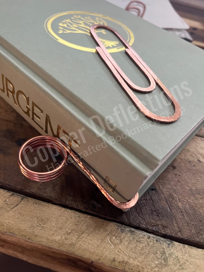 Puede incluir: Un marcap&aacute;ginas de cobre con forma de clip grande con un anillo de cobre m&aacute;s peque&ntilde;o unido a la parte inferior. El marcap&aacute;ginas est&aacute; encima de un libro verde con letras doradas. El texto del libro dice "Copper Defflections Handcrafted Bookmarks".