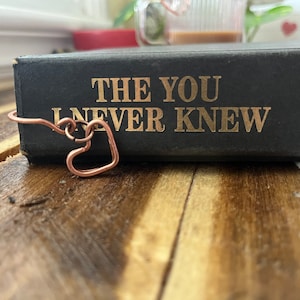 May include: A black book with the title "The You I Never Knew" on the spine. A copper wire bookmark shaped like a heart is on the book.