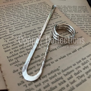 Puede incluir: Un marcap&aacute;ginas de plata con forma curva y superficie texturizada. El marcap&aacute;ginas est&aacute; sobre una p&aacute;gina de un libro con texto sobre el idioma delaware.