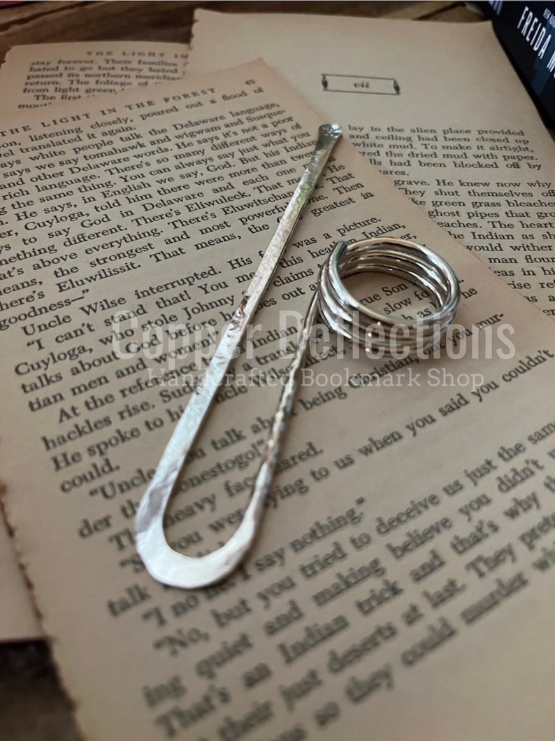 Puede incluir: Un marcap&aacute;ginas de plata con forma curva y superficie texturizada. El marcap&aacute;ginas est&aacute; sobre una p&aacute;gina de un libro con texto sobre el idioma delaware.