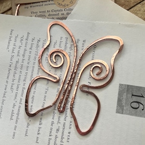 May include: A copper butterfly-shaped bookmark with a hammered texture. The bookmark is curled to hold a page in a book.