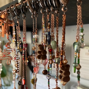 May include: A display of handmade keychains with copper-colored chains and clasps. Each keychain features a unique design, including colorful beads, heart-shaped charms, and small brown teddy bear pendants. The keychains are hanging from a dark wooden rod.