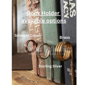 May include: Book holder made of wire in salvaged copper, sterling silver, and brass. The book holder is shown holding up a stack of books. The text "Book Holder available options" is visible at the top of the image. The text "Salvaged Copper", "Sterling Silver", and "Brass" are visible below the book holder.