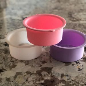 Könnte beinhalten: Drei Silikon-Backformen in verschiedenen Farben: weiß, rosa und lila. Die Formen sind rund und haben einen Rand oben.