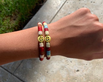 Può includere: Due braccialetti colorati con ciondoli a forma di faccina sorridente. I braccialetti sono realizzati con perline rosse, bianche, blu e marroni.
