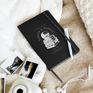 May include: Black notebook with the text "BORGIN & BURKES BOOKS" and a skull graphic. A light blue pen rests on the notebook, along with several small photographs and a camera. The notebook has a black elastic band closure and a ribbon bookmark.