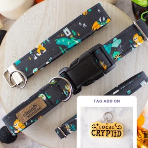 May include: A black fabric pet collar featuring a colorful pattern of forest creatures and trees, equipped with a black plastic buckle, a metal D-ring, and a brown rectangular brand tag. An inset shows a yellow tag with the text LOCAL CRYPTID.