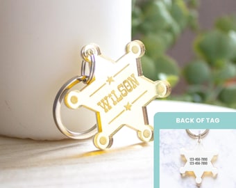 May include: A gold-toned acrylic star-shaped sheriff badge pet tag with a metal keyring. The front features the name WILSON engraved in the centre with small stars. An inset shows the back of the tag with the text 123-456-7890 printed twice.