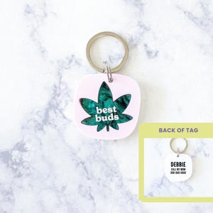 May include: A pink keychain with a green cannabis leaf design and the words "best buds" in white. The keychain has a silver ring and a square shape. The back of the tag is visible, with the text "DEBBIE CALL MY MOM 000-000-0000".