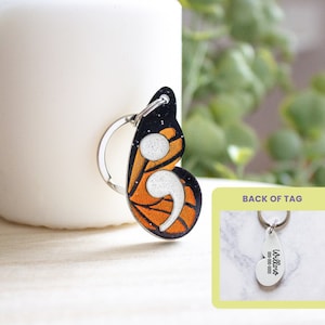 May include: A butterfly-shaped keychain with a semicolon design. The keychain features a monarch butterfly wing pattern in orange, black, and white. The back of the tag is visible, with the text "Willow" and a phone number.