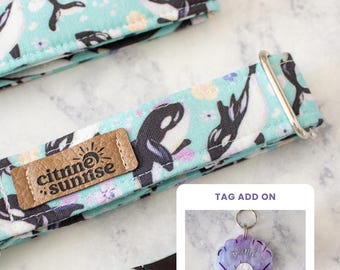 May include: A light blue fabric collar featuring a repeating pattern of black and white orca whales and small flowers. A tan leather tag with the text citrine sunrise is attached. An inset shows a purple seashell-shaped tag with a white pearl.