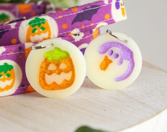 May include: A purple fabric collar with a Halloween pattern featuring pumpkins and bats, accompanied by two round white charms. One charm displays an orange glitter pumpkin, while the other features a purple glitter ghost design with metal rings.