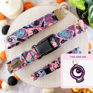May include: A set of pet accessories featuring a colorful tarot card design. The set includes a collar with a black buckle, a matching wristlet, and a small tag with a crystal ball graphic. The accessories have a purple, pink, and teal color scheme.