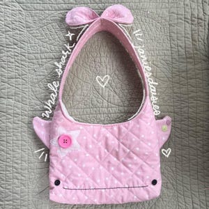 Può includere: Borsa a mano a forma di squalo balena trapuntata rosa con pois bianchi. Presenta un bottone a forma di stella, un dettaglio a fiocco e lati a forma di pinna. La borsa ha un manico e una bocca cucita. Il testo "whale shark" e "ljanesdaisies" sono scritti.