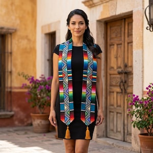 May include: A woman wearing a black dress and a colorful woven sash with a geometric pattern. The sash features blue, red, yellow, and green stripes, with gold tassels at the ends. The background includes a stone building and wooden doors.