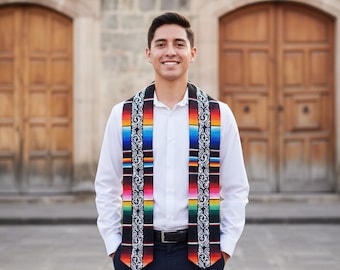 May include: A colorful graduation stole with a black border and a vibrant striped design in red, orange, yellow, green, and blue. The stole has a decorative pattern and gold tassels at the ends. The person is wearing a white shirt and dark pants.
