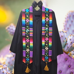 May include: Black graduation gown with a colorful embroidered sash featuring floral and striped patterns. The sash has a rainbow of colors, including red, orange, yellow, green, blue, and purple. Gold tassels hang from the bottom of the sash, and a black bow is at the top.