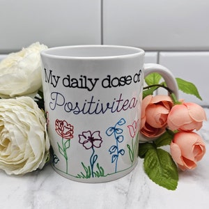 以下が含まれることがあります： 白いセラミック製のマグカップに、黒い文字で「My daily dose of Positivitea」と、カラフルな花柄が描かれています。
