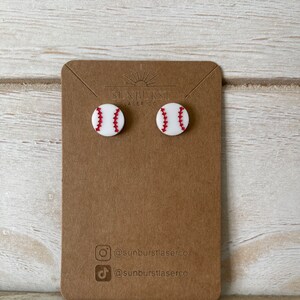 May include: A pair of white baseball earrings with red stitching. The earrings are displayed on a brown card with the text "Sunburst Laser Co." and social media handles.