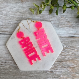 May include: A pair of pink acrylic earrings with the word "BRIDE" spelled out on each earring. The earrings are hanging from gold hooks.